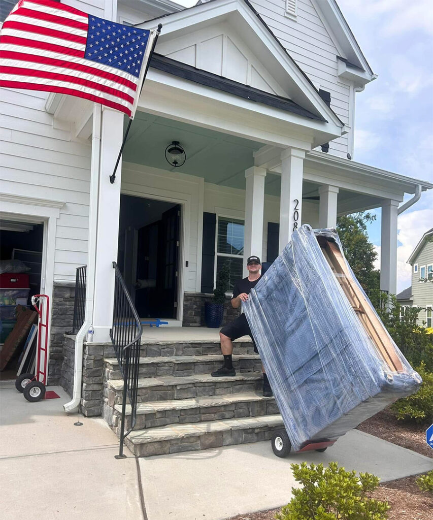 A professional mover using a hand truck to carefully move furniture down the front steps of a white suburban home with an American flag.